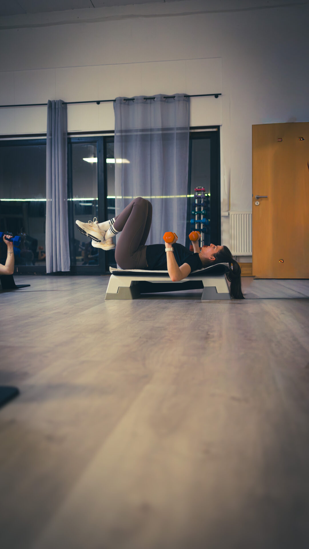 Eine Teilnehmerin macht eine Kraftübung auf einem Stepper mit angewinkelten Beinen und Freihanteln